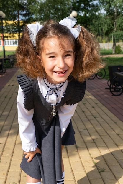 Chica alegre y divertida con una sonrisa desdentada en uniforme escolar con lazos blancos en el patio de la escuela Regreso a la escuela 1 de septiembre Un alumno feliz Clase primaria de educación primaria Retrato de un estudiante
