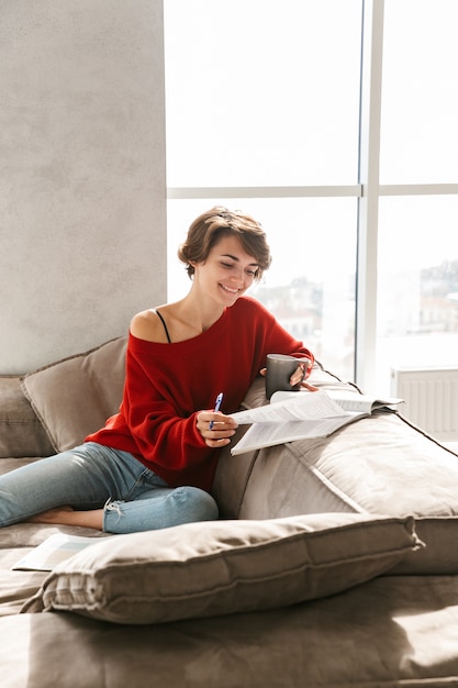 Foto chica alegre estudiando en casa mientras está acostado en un sofá y bebiendo té