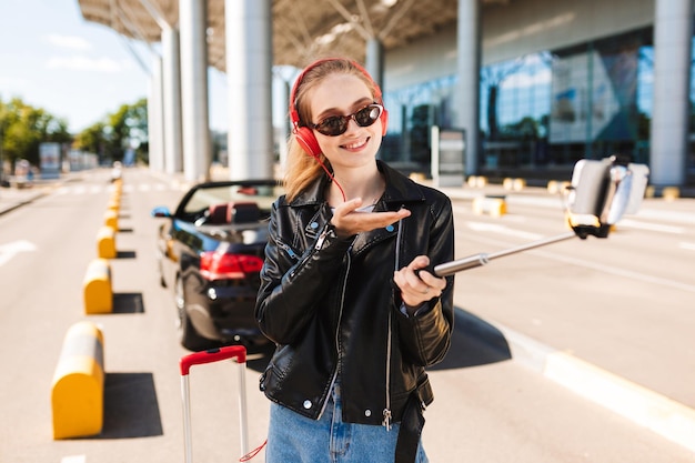 Chica alegre con gafas de sol y auriculares felizmente tomando fotos en el teléfono móvil con un auto descapotable negro en el fondo