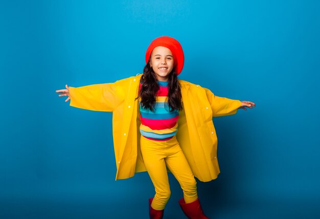 Una chica alegre con un impermeable amarillo, boina roja y botas de goma salta con los brazos extendidos y mira a la cámara.