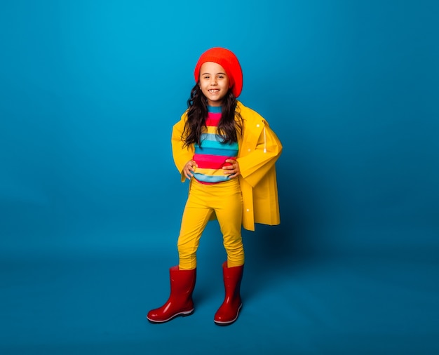 Una chica alegre con un impermeable amarillo, boina roja y botas de goma salta con los brazos extendidos y mira a la cámara.