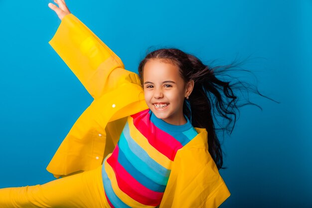 Una chica alegre con un impermeable amarillo y botas de goma rojas salta con los brazos extendidos y mira a la cámara.