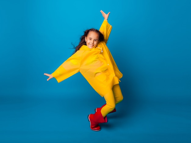 Una chica alegre con un impermeable amarillo y botas de goma rojas salta con los brazos extendidos y mira a la cámara.