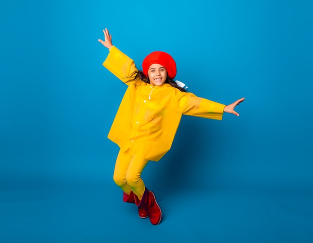 Una chica alegre con un impermeable amarillo y botas de goma rojas salta con los brazos extendidos y mira a la cámara.