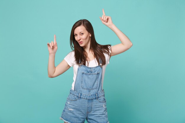 Chica alegre joven morena en ropa casual de mezclilla posando aislado en retrato de estudio de fondo de pared azul turquesa. Concepto de estilo de vida de personas. Burlarse del espacio de la copia. Señalar con los dedos índices hacia arriba.