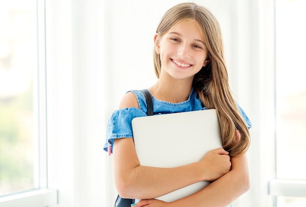 Chica alegre con mochila sosteniendo portátil en luz