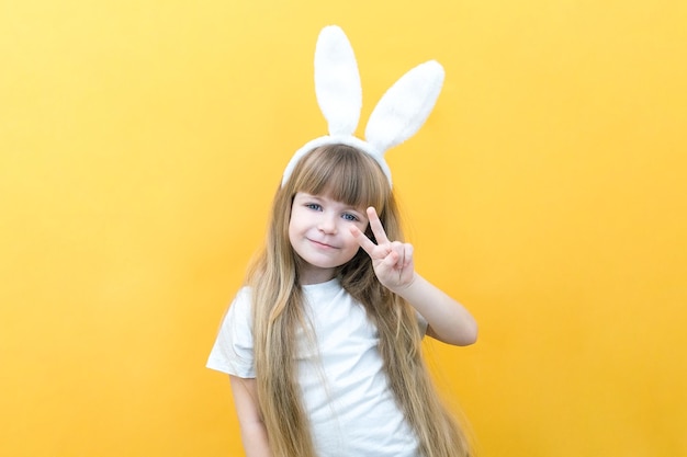 Chica alegre con orejas de conejo en la cabeza sobre un fondo amarillo Manos de niño feliz loco divertido como un conejo Preparación para el espacio de copia de vacaciones de Pascua para maqueta de texto