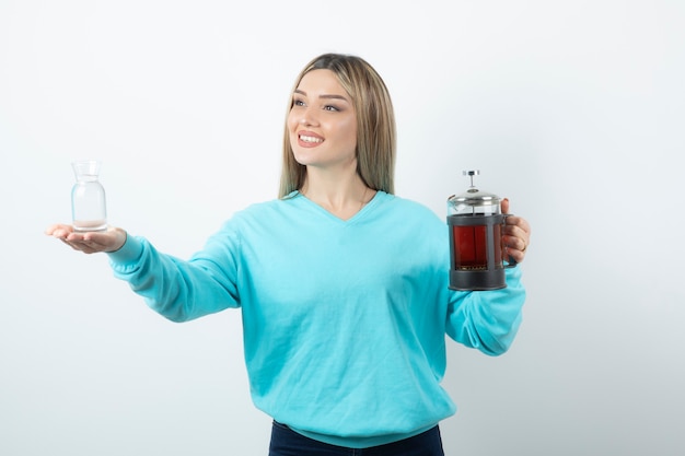 Foto chica alegre positiva sosteniendo la tetera con una taza de vidrio contra la pared blanca