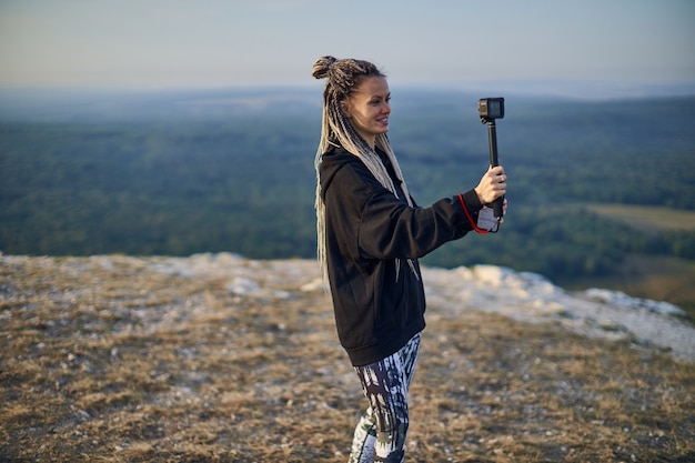 Foto una chica alegre con rastas en una chaqueta negra graba un blog en la cámara de pie en la cima de un monte ...