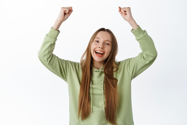 Chica alegre regocijándose de ganar y gritando de alegría, sí, levantando las manos animando al equipo a convertirse en ganador, lograr el objetivo y celebrar de pie sobre fondo blanco.