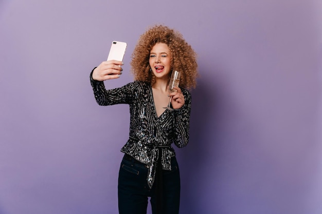 Chica alegre con rizos rubios muestra su lengua y hace selfie Retrato de chica en blusa brillante sosteniendo una copa de champán