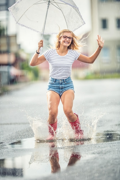 Chica alegre saltando con paraguas blanco en chanclos rojos punteados. Día caluroso de verano después de la lluvia mujer saltando y chapoteando en el charco