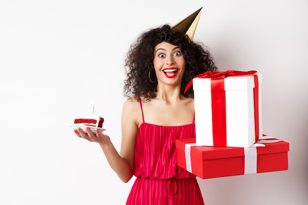 Chica alegre con sombrero de fiesta y vestido rojo, celebrando un cumpleaños, sosteniendo un regalo sorpresa y un b-day cake, disfrutando de las vacaciones, de pie sobre un fondo blanco.