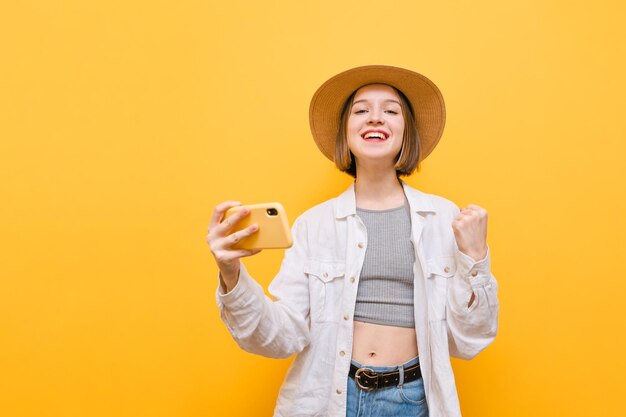 chica alegre con sombrero y ropa de verano sosteniendo un teléfono inteligente y mirando a la cámara