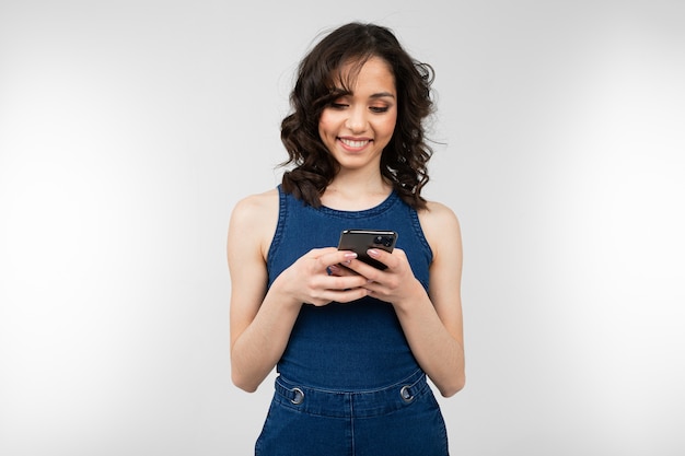 Chica alegre con una sonrisa en un vestido azul sostiene un teléfono inteligente y brilla de felicidad sobre un fondo blanco.