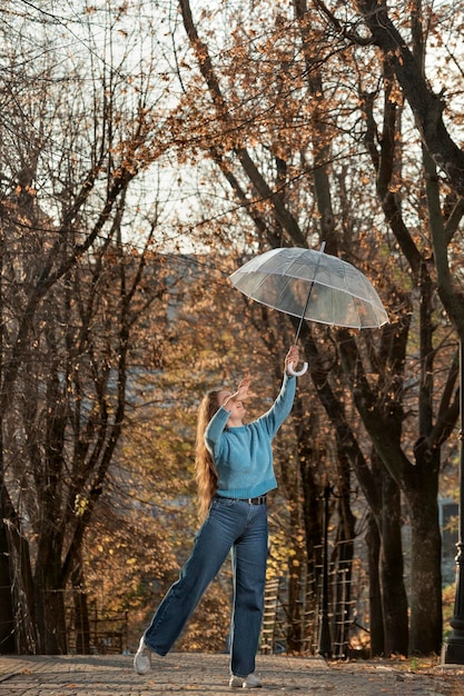 Chica alegre en suéter azul y pantalones con paraguas transparente en el callejón Baile de mujer hermosa en el parque de otoño