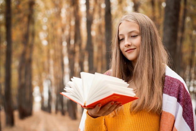 Chica alegre en suéter naranja y cuadros sobre sus hombros sosteniendo un libro en un parque de otoño