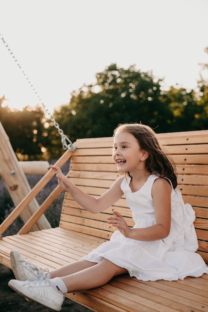 Chica alegre vestida con un vestido blanco está sentada en un columpio divirtiéndose en una noche de verano