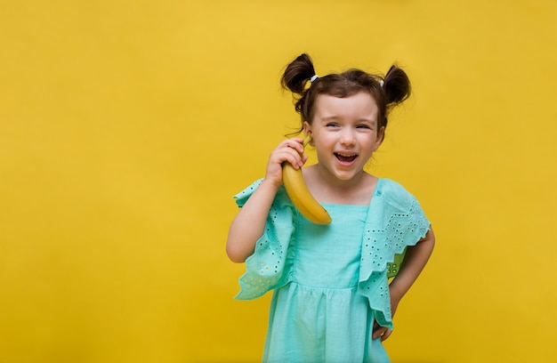 Chica alegre con un vestido azul se encuentra con un plátano y mira a la cámara sobre un fondo amarillo con una copia del espacio. Fondo de verano