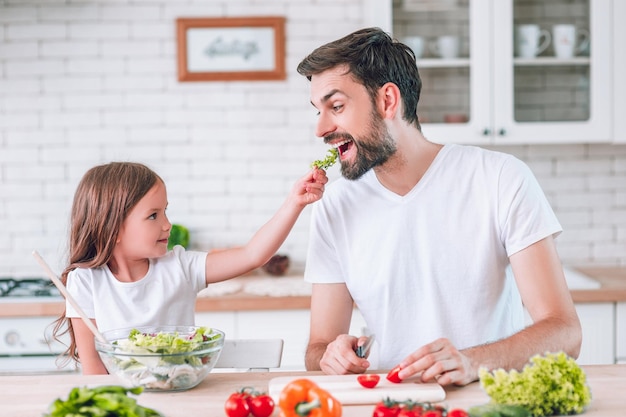 Chica alimentando a papá con hojas de lechuga mientras preparan ensalada en la cocina