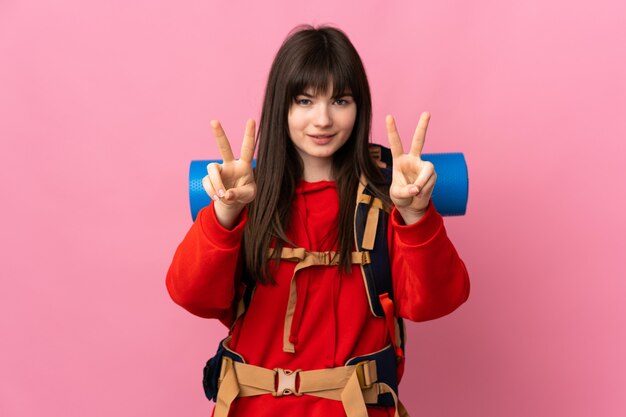 Chica alpinista con una mochila grande aislada en rosa sonriendo y mostrando el signo de la victoria