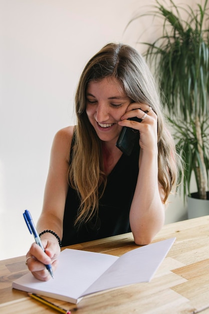 Chica alternativa joven contestando llamadas desde su teléfono celular y tomando notas en su sala de estar