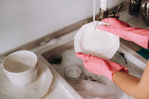 Chica ama de casa en guantes rosas lava platos a mano en el fregadero con detergente. La niña lava platos con guantes en casa.