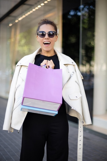 Foto chica americana riéndose con una camiseta negra y una chaqueta blanca y gafas de sol agarrando bolsas coloridas