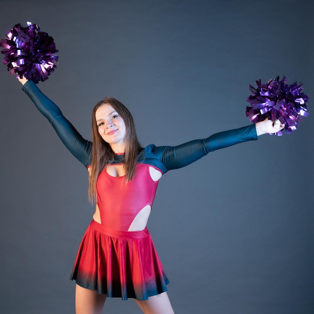 Chica animadora feliz en uniforme bailando con pompones aislado sobre fondo gris