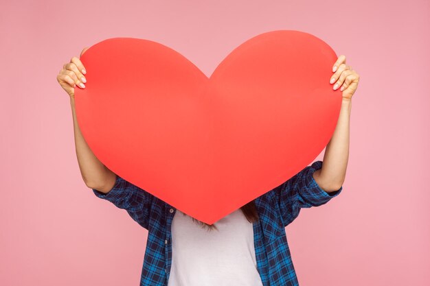 Chica anónima con pantalones a cuadros escondida detrás de un gran corazón de papel, sosteniendo una gran tarjeta de saludo y felicitando el día de San Valentín, expresando amor. tiro de estudio interior aislado sobre fondo de color rosa