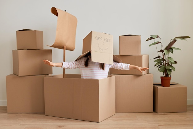 Chica anónima sentada rodeada de paquetes con cosas personales, posando con una caja de cartón con cara sonriente ahogada en lugar de cabeza, divirtiéndose durante la mudanza a un nuevo apartamento, extendiendo las manos