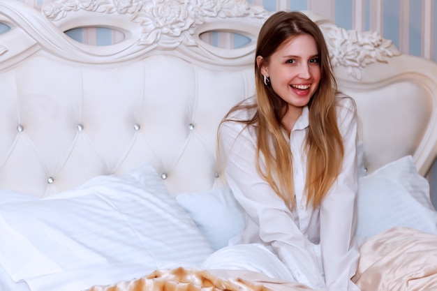 Chica de apariencia eslava en camisa sexy blanca en la cama en una habitación de hotel. Bonita mujer muestra las emociones del actor. Encantadora joven sentada en el sofá en el dormitorio y jugando con sus sentimientos. Relajarse concepto