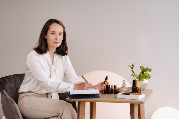 La chica aromaterapeuta está sentada en su oficina y sosteniendo una botella de aceite aromático en sus manos y escribiendo algo, hay aceites esenciales en la mesa