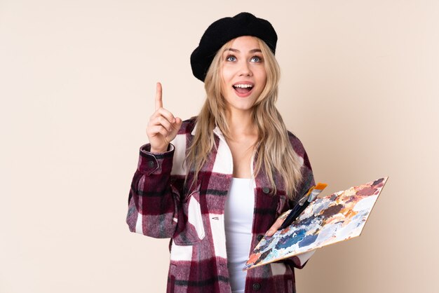 Chica artista adolescente sosteniendo una paleta en la pared azul apuntando hacia arriba y sorprendido