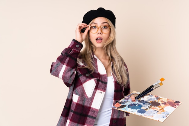 Foto chica artista adolescente sosteniendo una paleta en la pared azul con gafas y sorprendido