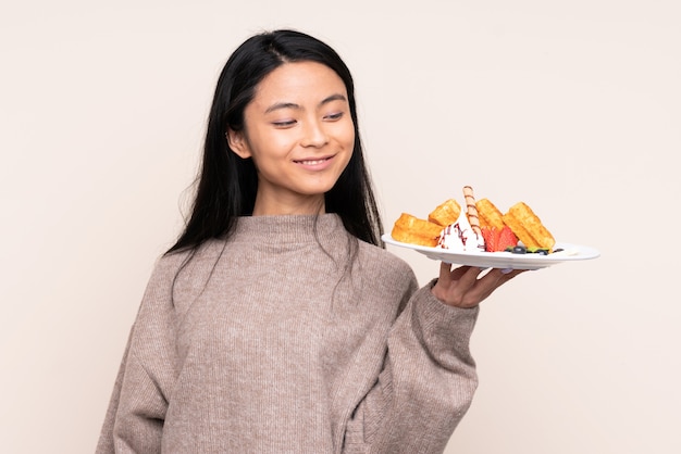 Chica asiática adolescente con gofres en la pared de color beige con expresión feliz