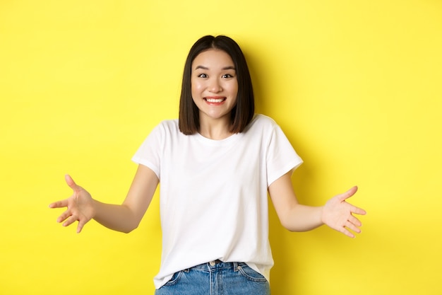 Chica asiática alegre sosteniendo algo grande y largo, sonriendo feliz a la cámara, de pie sobre amarillo.