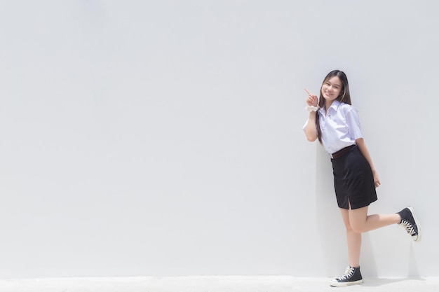 Una chica asiática con un alegre uniforme escolar sonríe y muestra su dedo para presentar algún producto