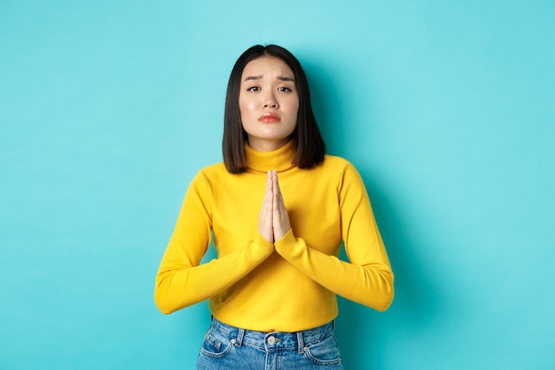 Chica asiática atractiva joven pidiendo un favor, necesita algo y pidiendo por favor, tomados de la mano en oración, de pie sobre fondo azul.