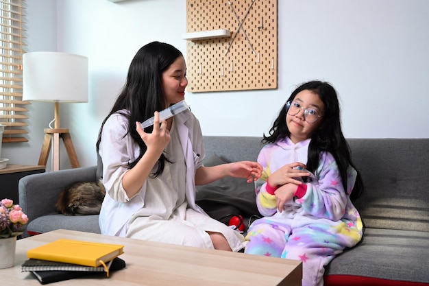 Chica asiática con bata blanca fingiendo ser médico vacunando a su hermana menor