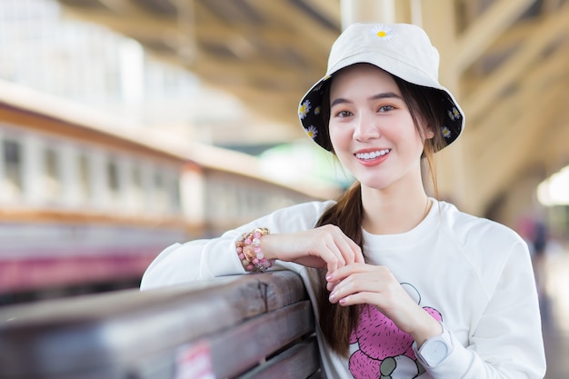 Chica asiática con una camisa blanca de manga larga se sienta feliz en la estación de tren esperando el tren