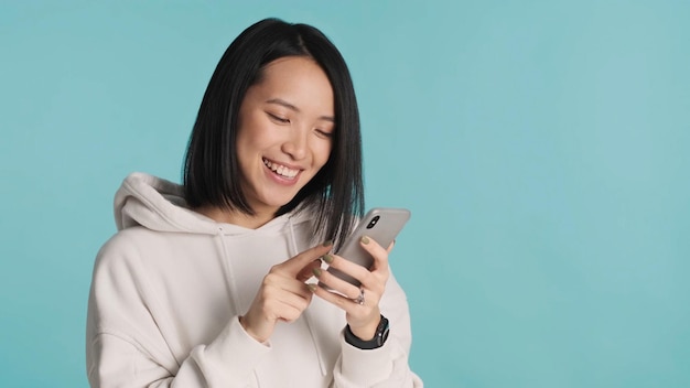 Chica asiática con capucha blanca enviando mensajes de texto con amigos y sonriendo usando un teléfono inteligente sobre un fondo colorido