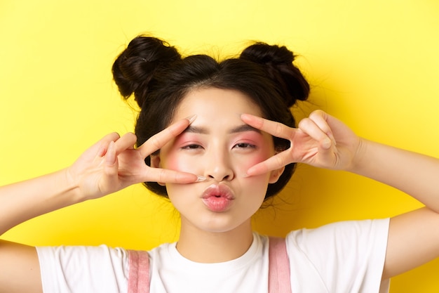 Foto chica asiática despreocupada mostrando signos v y besando los labios, de pie en amarillo