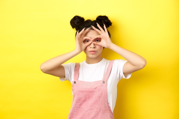 Chica asiática divertida mira a través de binoculares de mano, mirándote, de pie en ropa de verano en amarillo.