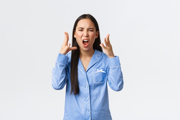 Chica asiática enojada enojada en pijama azul discutiendo, discutiendo con su novia durante la fiesta de pijamas, jurando o maldiciendo a alguien, dándose la mano enojada, de pie fondo blanco