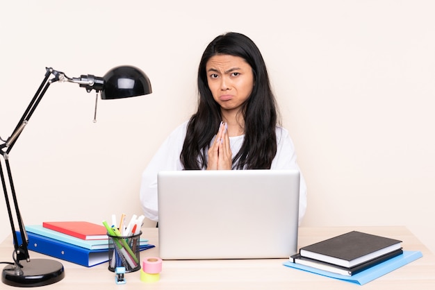 Chica asiática estudiante en un lugar de trabajo con una computadora portátil aislada en beige mantiene la palma unida. La persona pide algo
