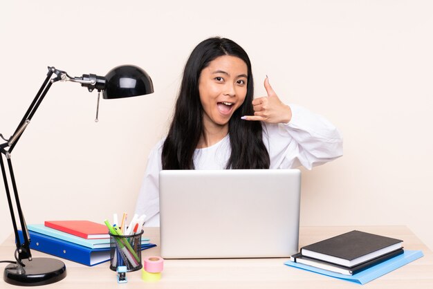 Chica asiática estudiante en un lugar de trabajo con una computadora portátil aislada en la pared beige haciendo gesto de teléfono. Llámame señal de vuelta