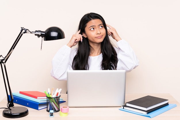 Chica asiática estudiante en un lugar de trabajo con una computadora portátil aislada sobre fondo beige con dudas y pensamiento