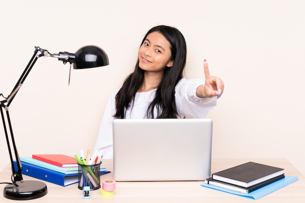 Chica asiática estudiante en un lugar de trabajo con una computadora portátil aislada sobre fondo beige mostrando y levantando un dedo