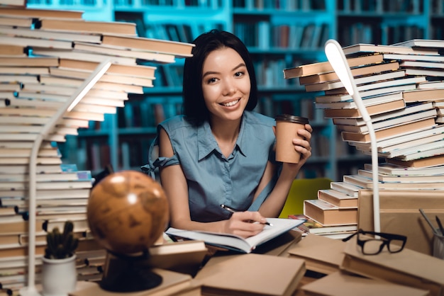 Foto chica asiática étnica rodeada de libros en la biblioteca. el estudiante está escribiendo en el cuaderno.